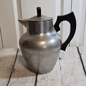 May include: A silver aluminum coffee pot with a black plastic handle. The pot has a textured surface and a rounded body.