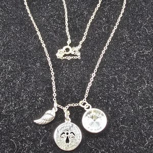 May include: Silver necklace featuring a wing charm, a circular pendant with an angel design, and a clear crystal. The necklace is presented on a black, textured background.
