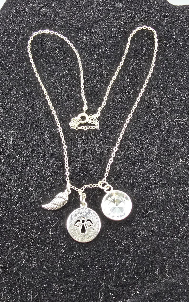 May include: Silver necklace featuring a wing charm, a circular pendant with an angel design, and a clear crystal. The necklace is presented on a black, textured background.