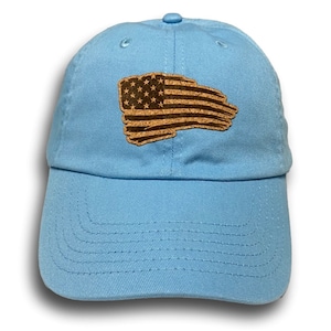 May include: Light blue baseball cap with a cork American flag patch. The cap has a curved brim and a fabric strap closure. The flag patch features stars and stripes in a distressed design.