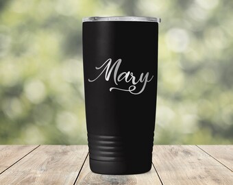 May include: A black insulated tumbler with a silver lid and the name "Mary" in cursive script. The tumbler has a cylindrical shape with a ridged bottom section. The background is a blurred green and brown.
