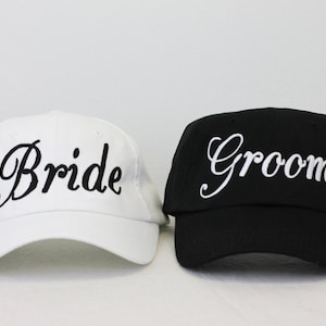 May include: Two baseball caps, one white with black embroidery that says "Bride" and one black with white embroidery that says "Groom".