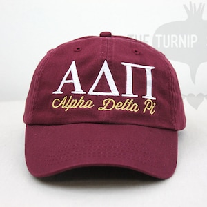 May include: A maroon baseball cap with white embroidered letters "AΔΠ" and gold embroidered text "Alpha Delta Pi".