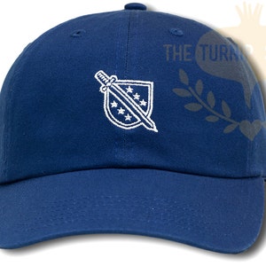 May include: A blue baseball cap with a white embroidered design of a sword and shield with stars. The text "THE TURN SEED CO." is visible in the background.
