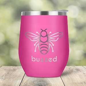 May include: A pink insulated wine tumbler with a silver lid. The tumbler features a silver outline of a bee and the word "buzzed" in silver lettering. The tumbler is designed to keep beverages cold.