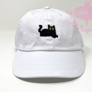May include: White baseball cap with a black embroidered cat with yellow eyes.