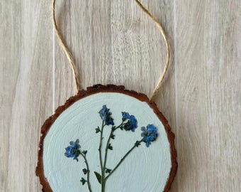 May include: A round wooden slice with a light blue painted surface, featuring pressed blue flowers. The wood has a natural bark edge and a twine loop for hanging. A rustic, nature-inspired decorative item.