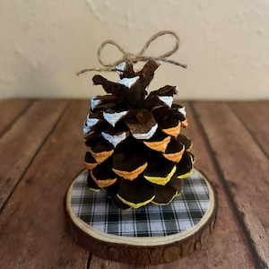 May include: A painted pine cone with orange, yellow, and white stripes sits on a small wooden slice with a black and white checkered pattern. The pine cone has a twine bow tied around the top.