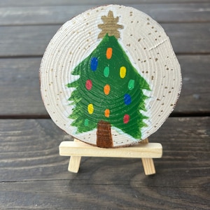 Könnte beinhalten: Ein handbemaltes Weihnachtsbaumornament aus Holz auf einem kleinen Staffelei. Der Baum ist grün mit einem braunen Stamm und bunten Ornamenten. Der Hintergrund ist weiß mit goldenem Glitzer.