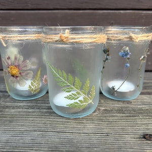 Puede incluir: Tres portavelas de cristal con flores y hojas secas. Cada tarro está decorado con un diseño botánico diferente, incluyendo una margarita, un helecho y flores azules. Los bordes están envueltos con hilo de cáñamo, y cada tarro contiene una vela de té encendida.