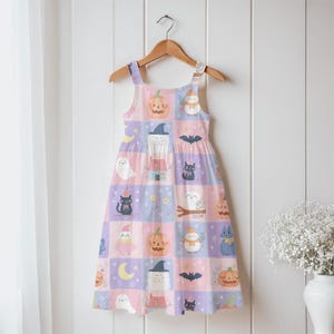K&ouml;nnte beinhalten: Ein &auml;rmelloses Kleid mit einem Karomuster aus pastellrosa und hellvioletten Quadraten. Der Druck zeigt Illustrationen von orangefarbenen K&uuml;rbissen, schwarzen Katzen, wei&szlig;en Geistern, Flederm&auml;usen, gelben Mondsicheln und kleinen H&auml;usern.