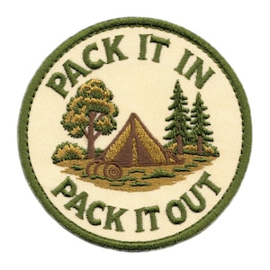 May include: A circular embroidered patch with a cream background and a dark green border. The design features a brown tent, a deciduous tree, and two pine trees. The text "PACK IT IN" and "PACK IT OUT" is embroidered in dark green lettering.