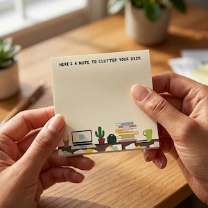 Op de afbeelding: Een vierkante, crèmekleurige plakbriefje, vastgehouden door handen. De notitie heeft de tekst "HERE'S A NOTE TO CLUTTER YOUR DESK." en een decoratieve rand met een laptop, cactus, boeken en een mok. De achtergrond is een houten bureau.