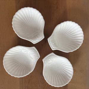 May include: Four white ceramic shell-shaped dishes arranged on a wooden surface. Each dish has a detailed, ridged design and a small leaf-like accent at the base. The dishes are empty and ready for use.
