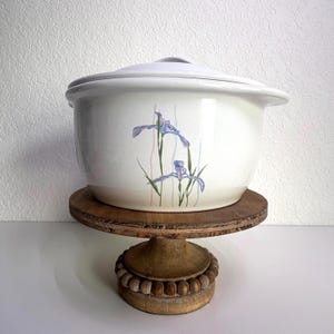 May include: A white ceramic casserole dish with a lid, featuring a floral iris design in shades of purple and green. The dish is displayed on a rustic, wooden pedestal with a round top and beaded base. A vintage kitchenware item.