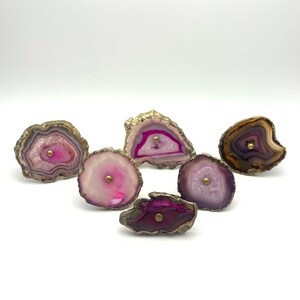 May include: A collection of six irregular agate stone drawer knobs featuring natural banded patterns in shades of pink, purple, and brown. Each stone slice is edged in a metallic gold finish and includes a centered brass-toned metal mounting bolt.