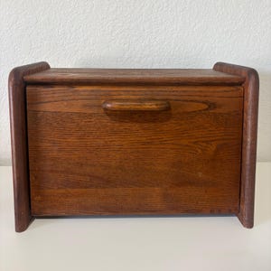 May include: A rectangular wooden bread box with a medium brown finish and visible wood grain. The unit features a front-hinged door with a horizontal wooden handle centered on the panel, framed by two curved side supports and a flat top surface.
