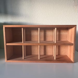 May include: A rectangular wooden storage shelf featuring a natural light brown finish. The unit is divided into two horizontal levels by a central shelf, with six vertical cylindrical wooden dowels positioned to create individual compartments.