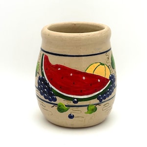 May include: A beige ceramic mug featuring a hand-painted design of a red watermelon slice, a yellow melon, and clusters of dark blue grapes. The artwork is framed by thin dark blue horizontal lines and accented with small green leaves and vines.