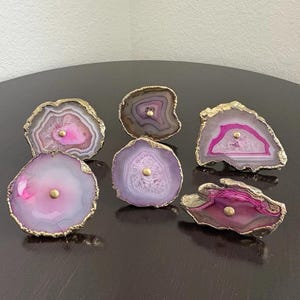 May include: A collection of seven agate cabinet knobs with gold-colored edges and a central gold-colored screw. The knobs feature a variety of colors, including pink, purple, and gray, and are designed to add a natural, elegant touch to furniture.