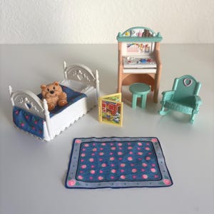 May include: A miniature dollhouse bedroom set with a white bed, a teddy bear, a desk, a rocking chair, a small table, a rug, and a book. The bed has a blue floral patterned blanket. The desk is light brown and green.