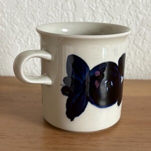 May include: Cream-colored ceramic mug with a curved handle. The mug features a dark blue floral design with pink accents. The mug is sitting on a wooden surface.
