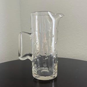May include: A clear glass pitcher with a handle and a spout. The pitcher has a textured surface with small, rounded bumps. The pitcher is sitting on a dark surface. The pitcher is likely used for serving beverages.