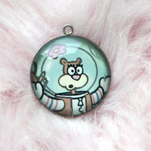 Puede incluir: Un colgante redondo con un personaje de dibujos animados con un traje espacial. El colgante tiene un fondo azul claro con una flor y la cara del personaje. El colgante está sujeto a un lazo plateado.