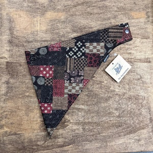 May include: A patchwork dog bandana with a variety of patterns in black, brown, and burgundy. The bandana has a snap closure and a tag that reads "the dog bandana."