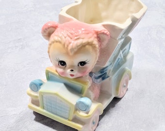 May include: A vintage ceramic planter shaped like a car, featuring a pink teddy bear driver. The car is painted in pastel colors, including blue, yellow, and pink. The planter has a heart-shaped opening.