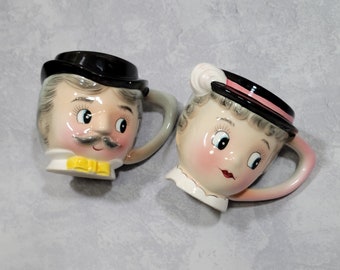 May include: Two ceramic mugs shaped like vintage faces, a man and a woman. The man mug has a black hat, mustache, and yellow bow tie. The woman mug has a black hat with a pink band and a white collar. Both mugs have pink cheeks and matching handles.