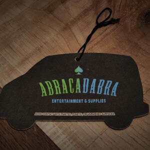 May include: Black car air freshener with the text "ABRACADABRA ENTERTAINMENT & SUPPLIES" and a green and blue spade symbol.
