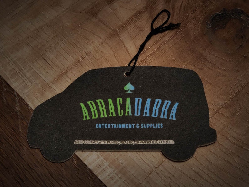 May include: Black car air freshener with the text "ABRACADABRA ENTERTAINMENT & SUPPLIES" and a green and blue spade symbol.