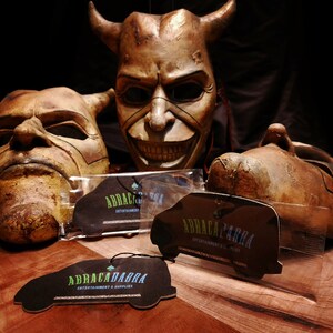 May include: Three brown, demonic-looking masks with horns and exaggerated features.  Three black car air fresheners with the text "ABRACADABRA ENTERTAINMENT & SUPPLIES" in green and blue lettering.
