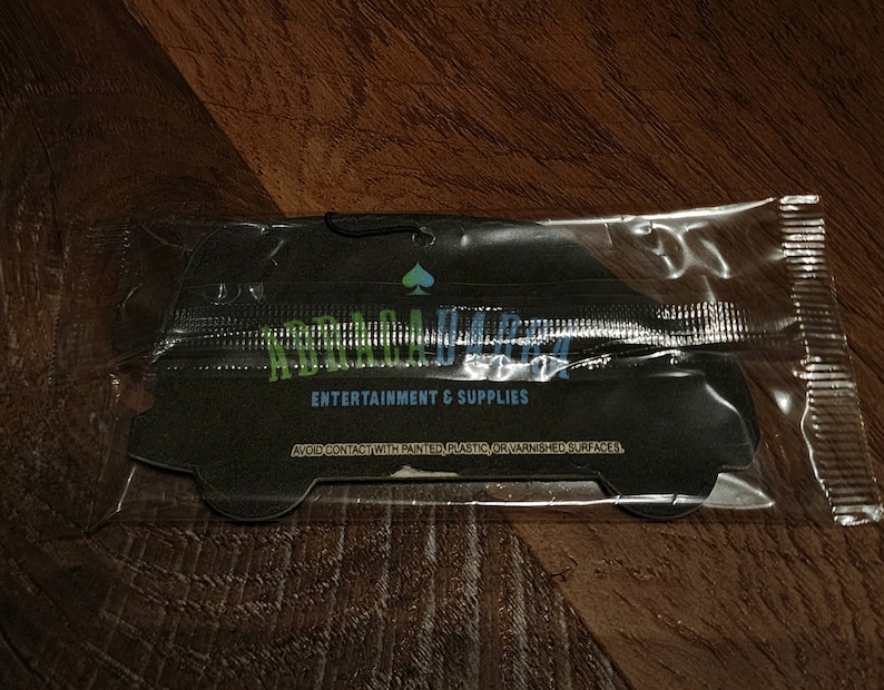 May include: A black car air freshener with the text "Remarkable" in green and blue letters. The text "Entertainment & Supplies" is printed below the logo. The air freshener is packaged in a clear plastic bag.