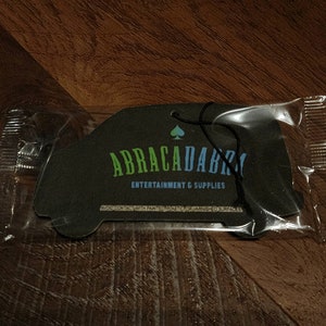 May include: A black car-shaped air freshener with the text "ABRACADABRA ENTERTAINMENT & SUPPLIES" and a spade symbol. The air freshener is packaged in a clear plastic bag.