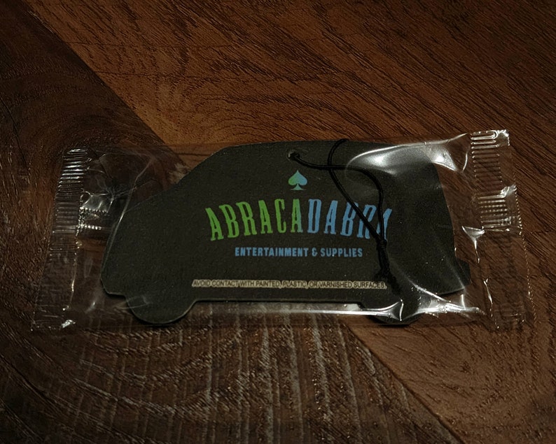 May include: A black car-shaped air freshener with the text "ABRACADABRA ENTERTAINMENT & SUPPLIES" and a spade symbol. The air freshener is packaged in a clear plastic bag.