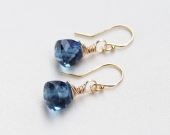 Könnte beinhalten: Ein Paar Ohrhänger mit facettierten, tropfenförmigen blauen Edelsteinen, die an goldfarbenen Drahtbügeln hängen. Jeder Stein ist oben mit einer feinen goldfarbenen Drahtwicklung befestigt, was ein schlichtes Design ergibt.