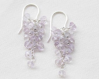 Könnte beinhalten: Ein Paar Ohrhänger mit Clustern aus kleinen, facettierten, hellvioletten Amethystperlen, die an silberfarbenen Metallhaken hängen. Die Perlen sind in einer kaskadierenden, traubenartigen Formation angeordnet und erzeugen ein strukturiertes Aussehen.