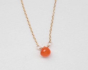 Könnte beinhalten: Eine zarte goldene Halskette mit einem einzelnen, kleinen, facettierten, tropfenförmigen Anhänger aus orangefarbenem Karneol. Der Stein ist mit einer kleinen, gewickelten Golddrahtschlaufe mittig an der dünnen Metallkette befestigt.