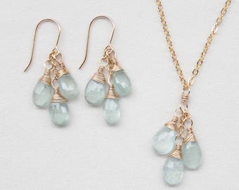 Könnte beinhalten: Ein Schmuckset bestehend aus einer Halskette und einem Paar Ohrringen. Jedes Stück zeigt mehrere blassblaue, tropfenförmige Edelsteine, die mit goldfarbenem Draht umwickelt sind. Die Halskette hat eine zarte goldene Kette.