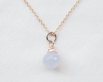 Könnte beinhalten: Eine zarte Halskette mit einer dünnen goldfarbenen Kette und einem einzelnen facettierten, hellblauen tropfenförmigen Edelsteinanhänger. Der Stein ist mit einem kleinen goldfarbenen Ring an der Kette befestigt und mit feinem Draht umwickelt.