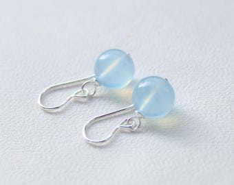 Könnte beinhalten: Ein Paar Ohrhänger mit glatten, runden, durchscheinenden hellblauen Perlen, die an einfachen silberfarbenen Metallhaken hängen. Die Perlen haben ein weiches, milchiges Aussehen und sind mit kleinen silbernen Ringen an den gebogenen Haken befestigt.