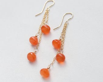 Könnte beinhalten: Ein Paar goldfarbene Ohrhänger mit zarten Ketten, die an geschwungenen Ohrhaken befestigt sind. Jeder Ohrring zeigt drei facettierte, runde, durchscheinende orangefarbene Karneolperlen, die in unterschiedlichen Längen an den Ketten hängen.
