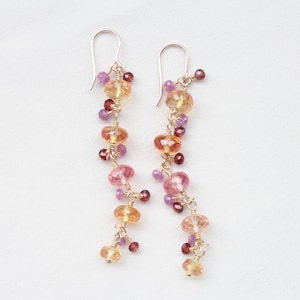 Puede incluir: Pendientes colgantes con una cascada de piedras preciosas de colores. Los pendientes presentan una mezcla de cuentas rosas, naranjas y moradas, ensartadas con alambre dorado. El diseño incluye pequeñas cuentas redondas y piedras más grandes facetadas.