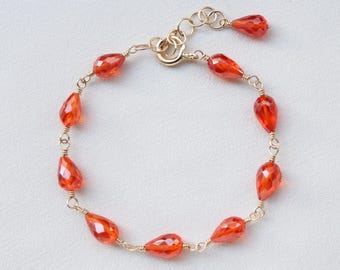Könnte beinhalten: Ein goldenes Kettenarmband mit mehreren facettierten, tropfenförmigen orangefarbenen Perlen. Das Armband besteht aus dünnen Golddrahtgliedern, die jede Perle verbinden, und ist mit einem runden Verschluss sowie einer zusätzlichen hängenden Perle versehen.