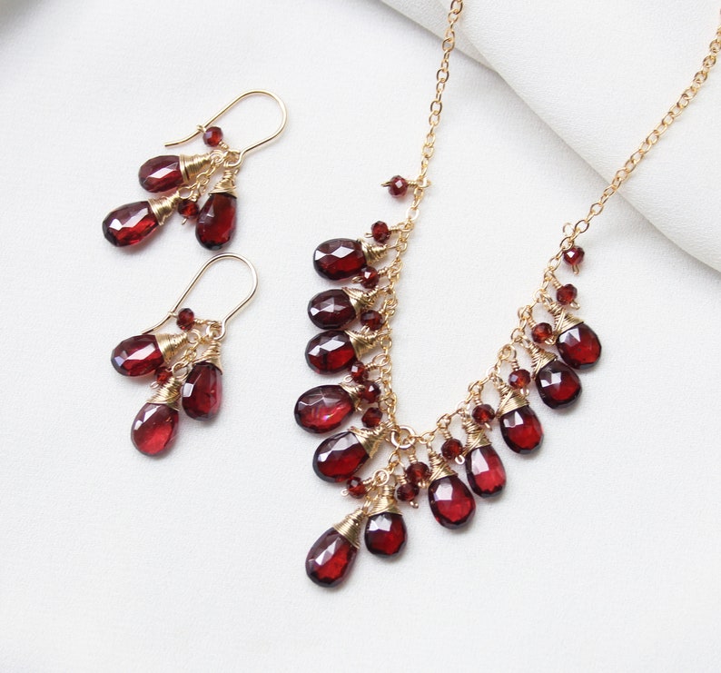 K&ouml;nnte beinhalten: Ein goldenes Halsketten- und Ohrring-Set mit baumelnden roten Granatsteinen. Die Halskette hat ein kaskadierendes Design mit mehreren Granattropfen.