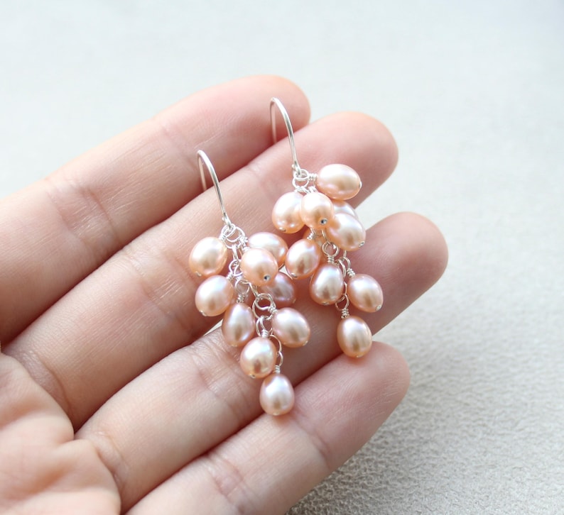 May include: A pair of dangle earrings with a cluster of blush pink freshwater pearls hanging from silver hooks.