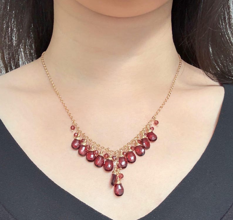 K&ouml;nnte beinhalten: Eine goldene Kettenkette mit einem tropfenf&ouml;rmigen Anh&auml;nger aus roten Edelsteinen. Die Kette hat ein kaskadierendes Design mit kleineren roten Edelsteinen, die an der Kette h&auml;ngen.