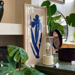 Pode incluir: Uma impress&atilde;o emoldurada de um l&iacute;rio azul sobre um fundo branco. A impress&atilde;o est&aacute; pendurada em uma parede ao lado de uma l&acirc;mpada com um abajur de vidro transparente e uma base branca.
